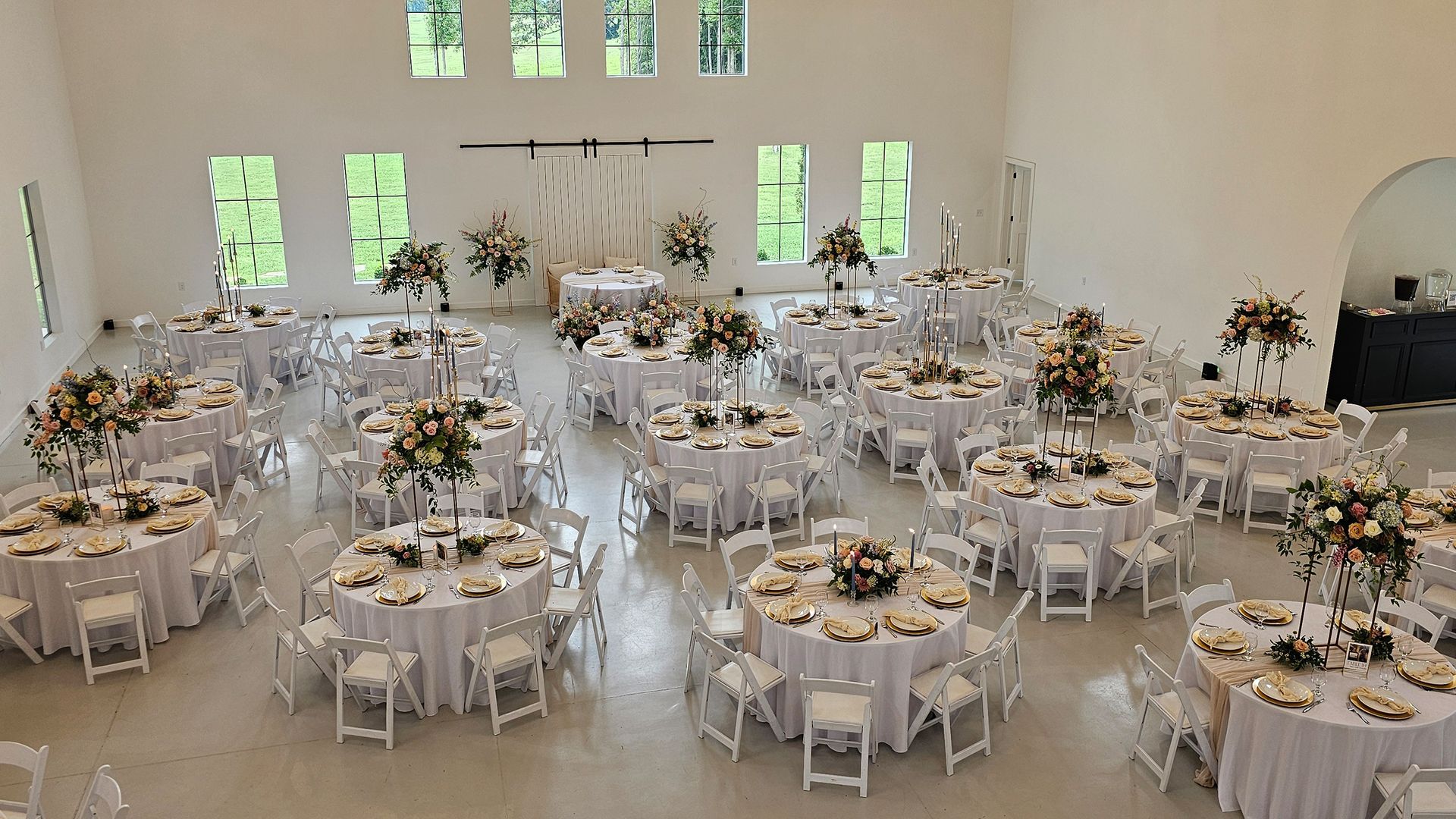 Wedding reception hall with round tables set for guests. White decor, floral centerpieces.
