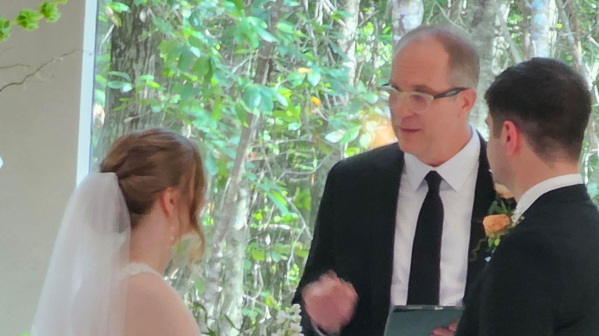Wedding ceremony with officiant, bride, and groom. Sunlight streams through trees in the background.