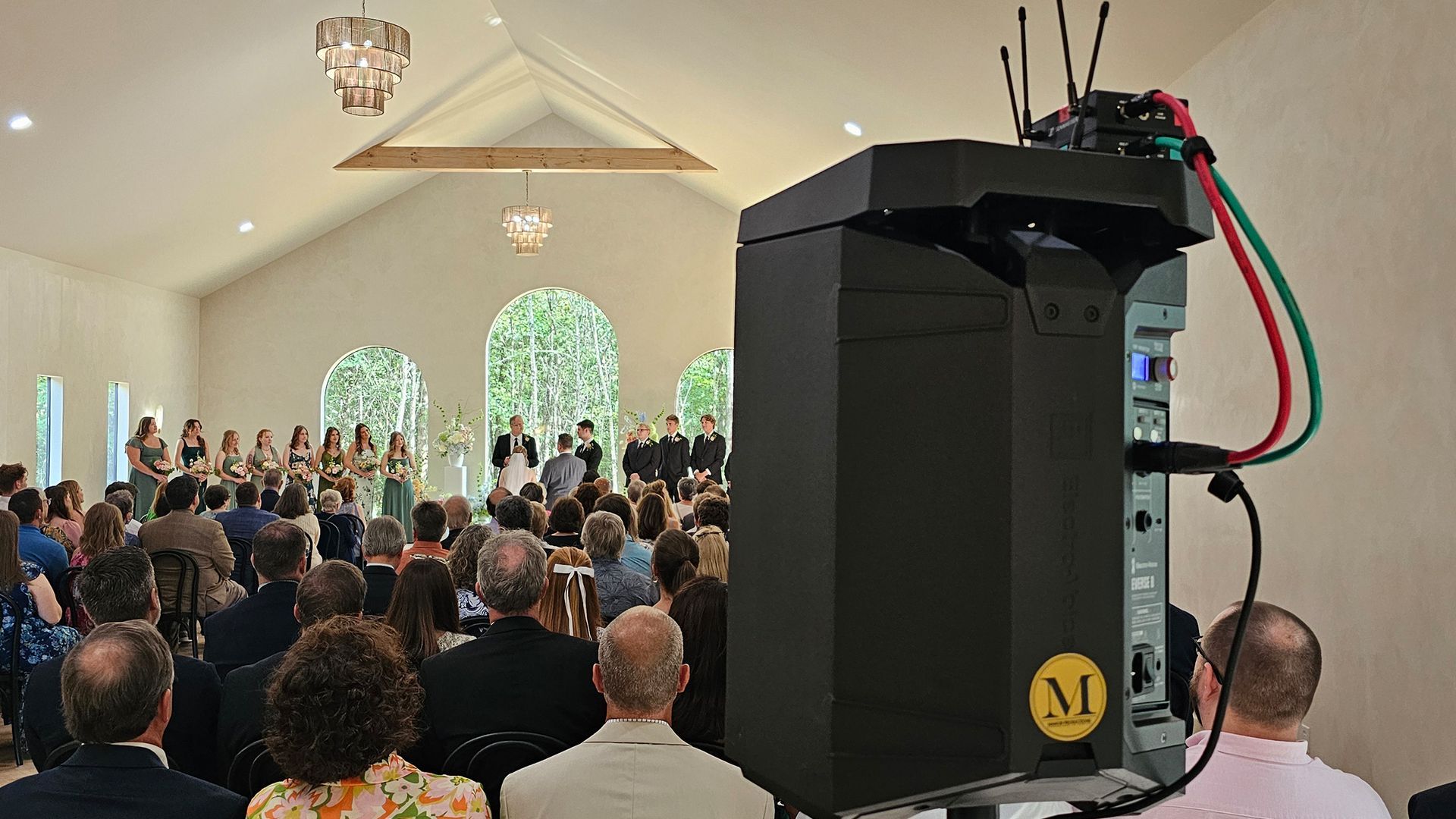 Wedding ceremony in a light-filled venue with a sound system in the foreground and guests watching.
