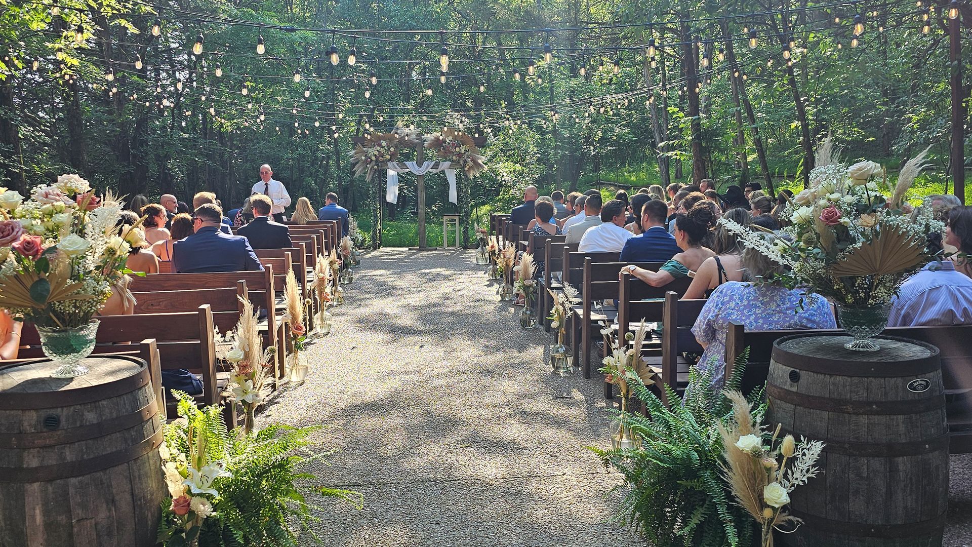 Wedding ceremony outdoors with guests seated on wooden benches, floral decorations, and an officiant at the front.