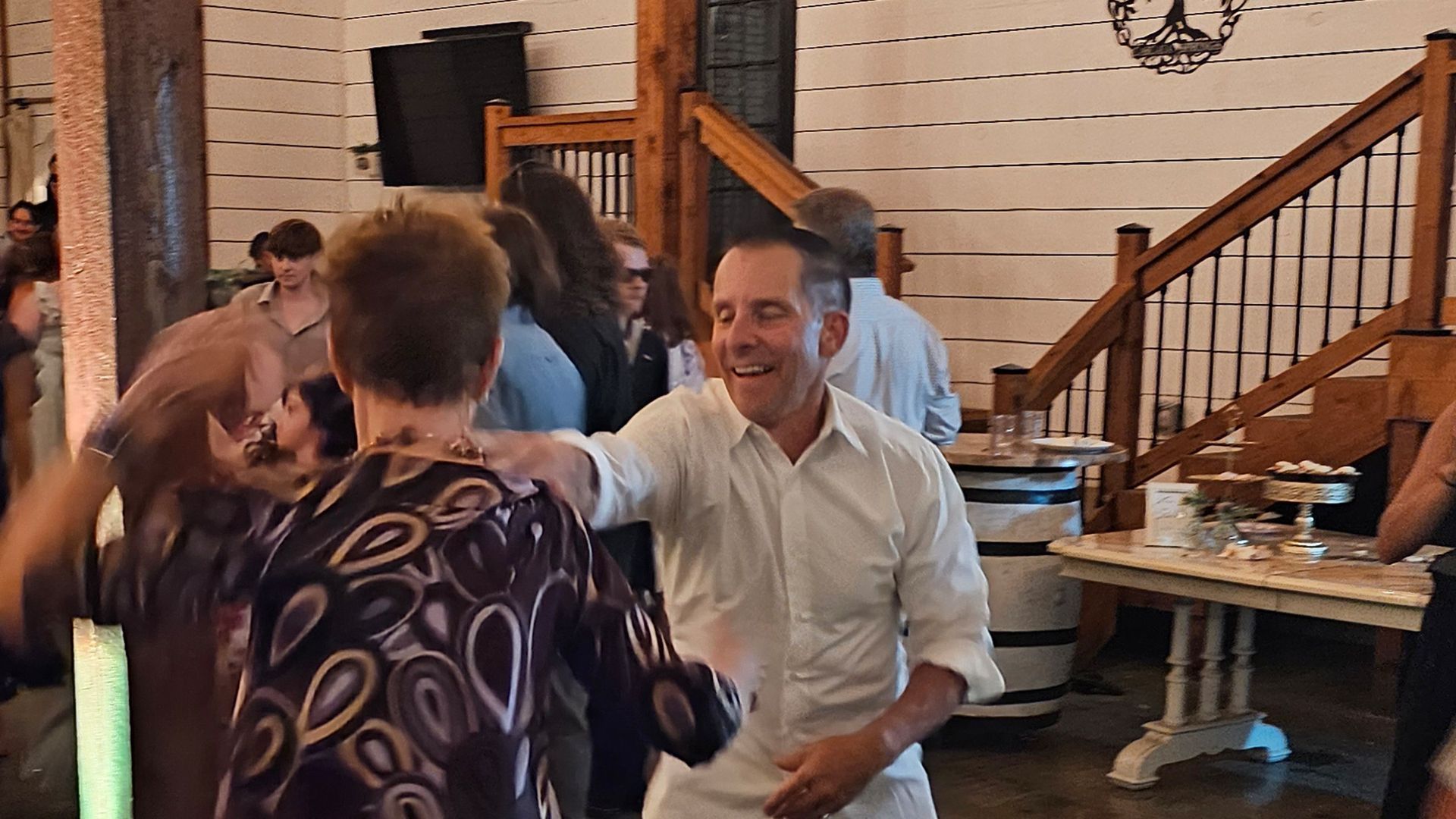 People dancing at an event in a rustic setting, man in white shirt smiles, wooden stairs in background.