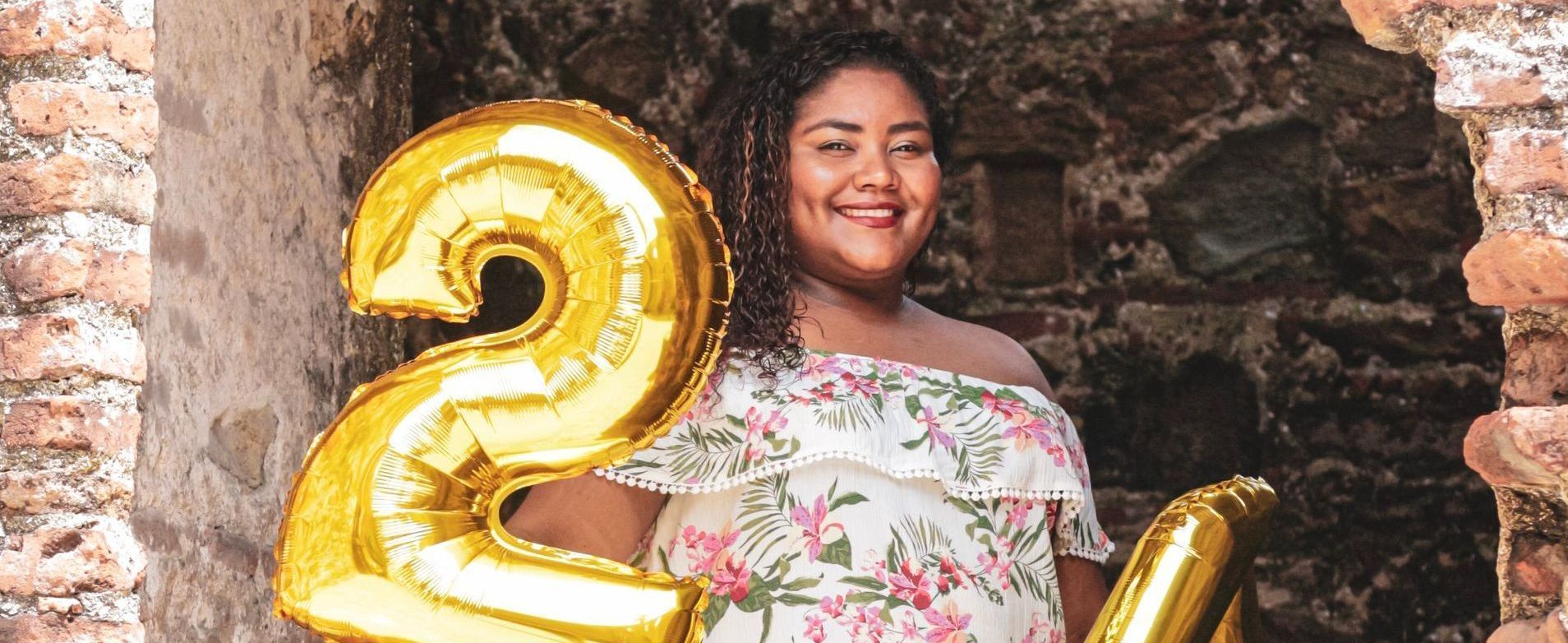 Woman holding gold balloon with the number 20, smiling, wearing floral top, brick wall background.