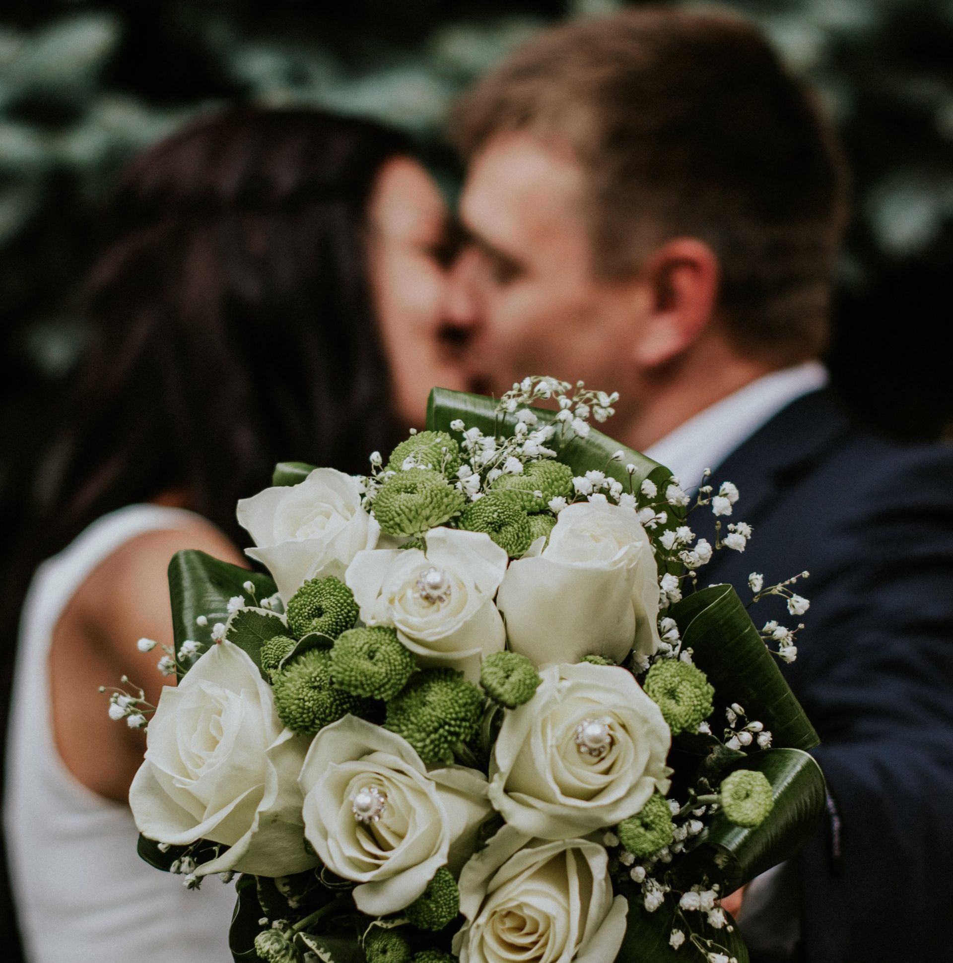 Wedding bouquet with white roses in focus, blurred couple kissing in the background.