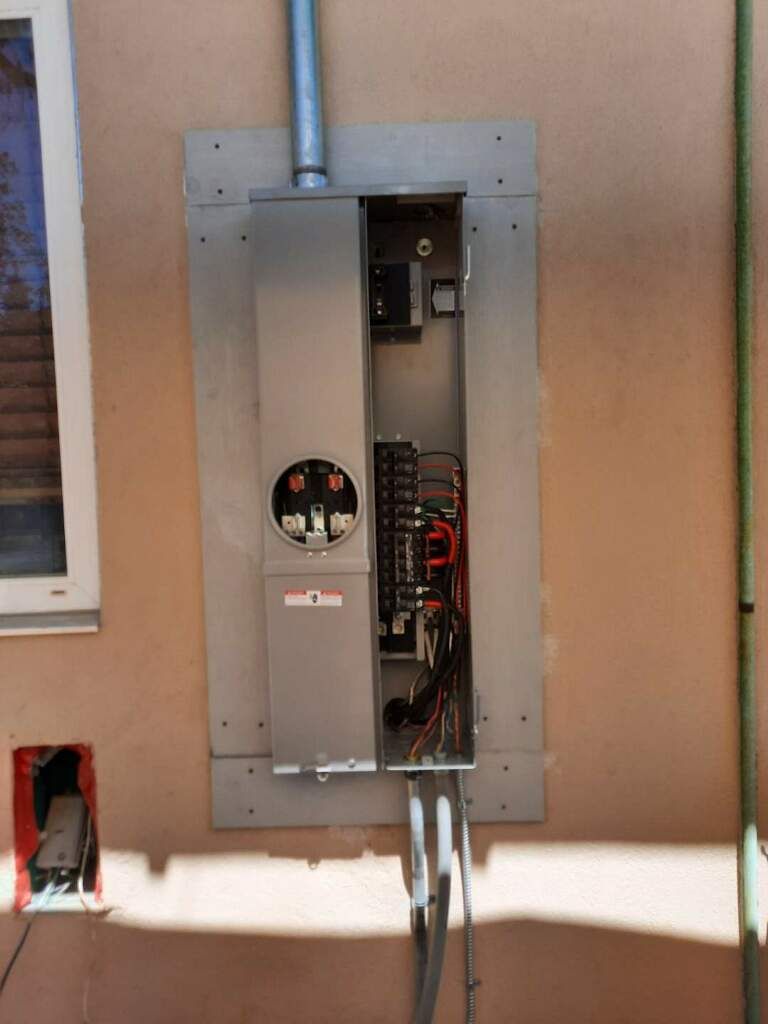 Open electrical service panel mounted on an exterior wall with wires exposed and conduit above.