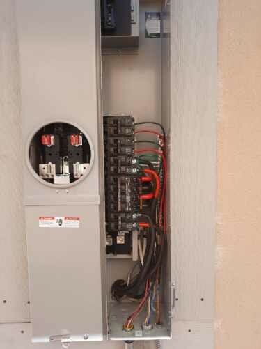 Open electrical panel with exposed wiring, breakers, and red cables in a utility room.