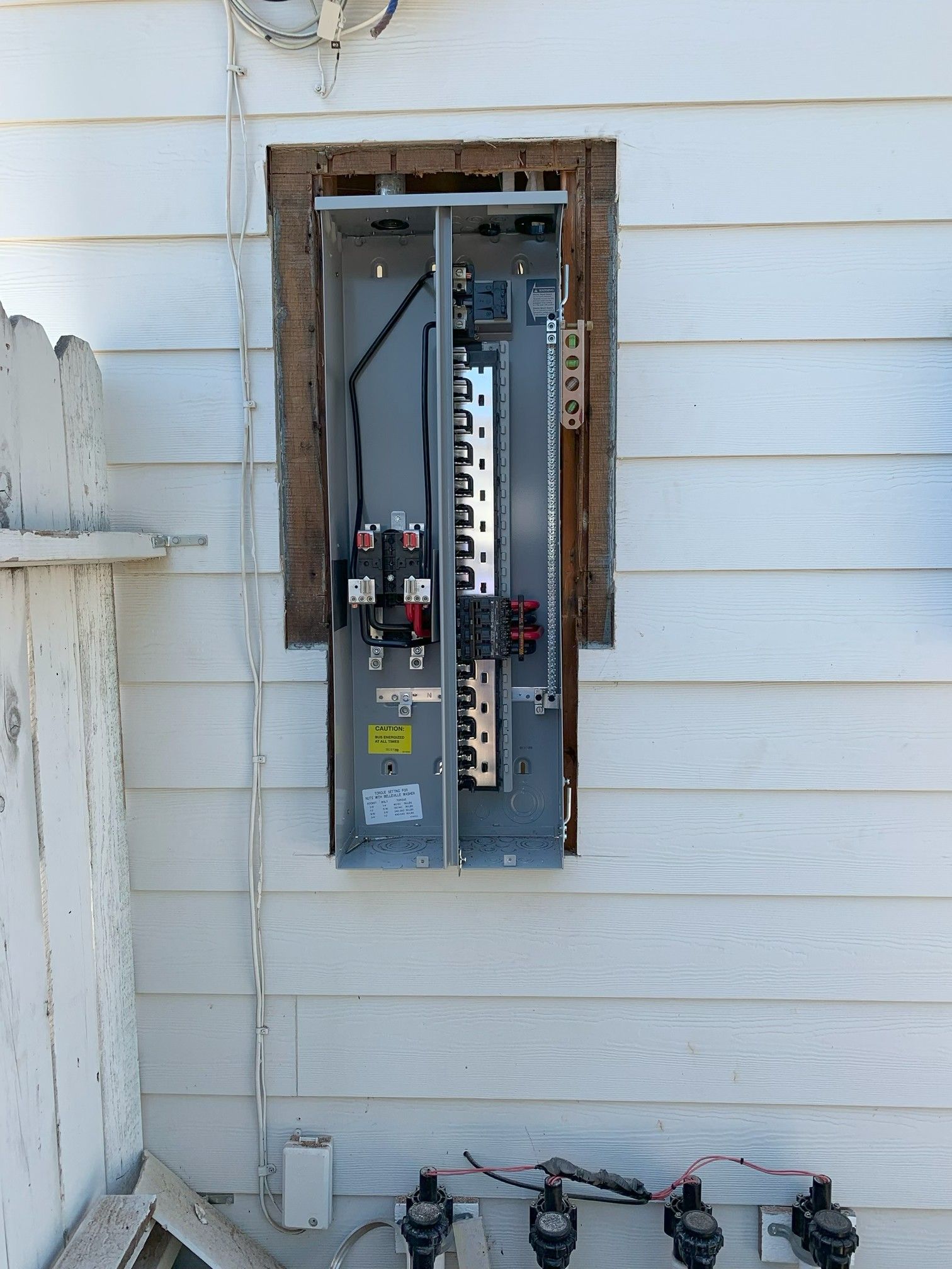Open electrical breaker panel mounted on a house exterior wall, with exposed wiring and piping below.