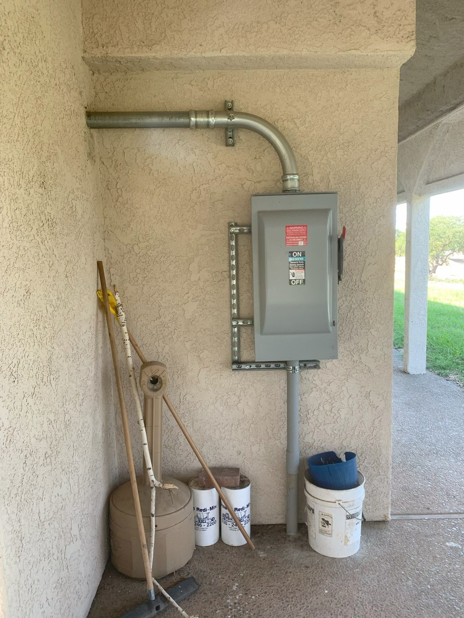 Gray electrical panel on a stucco wall with conduit, cleaning tools, and buckets beside it