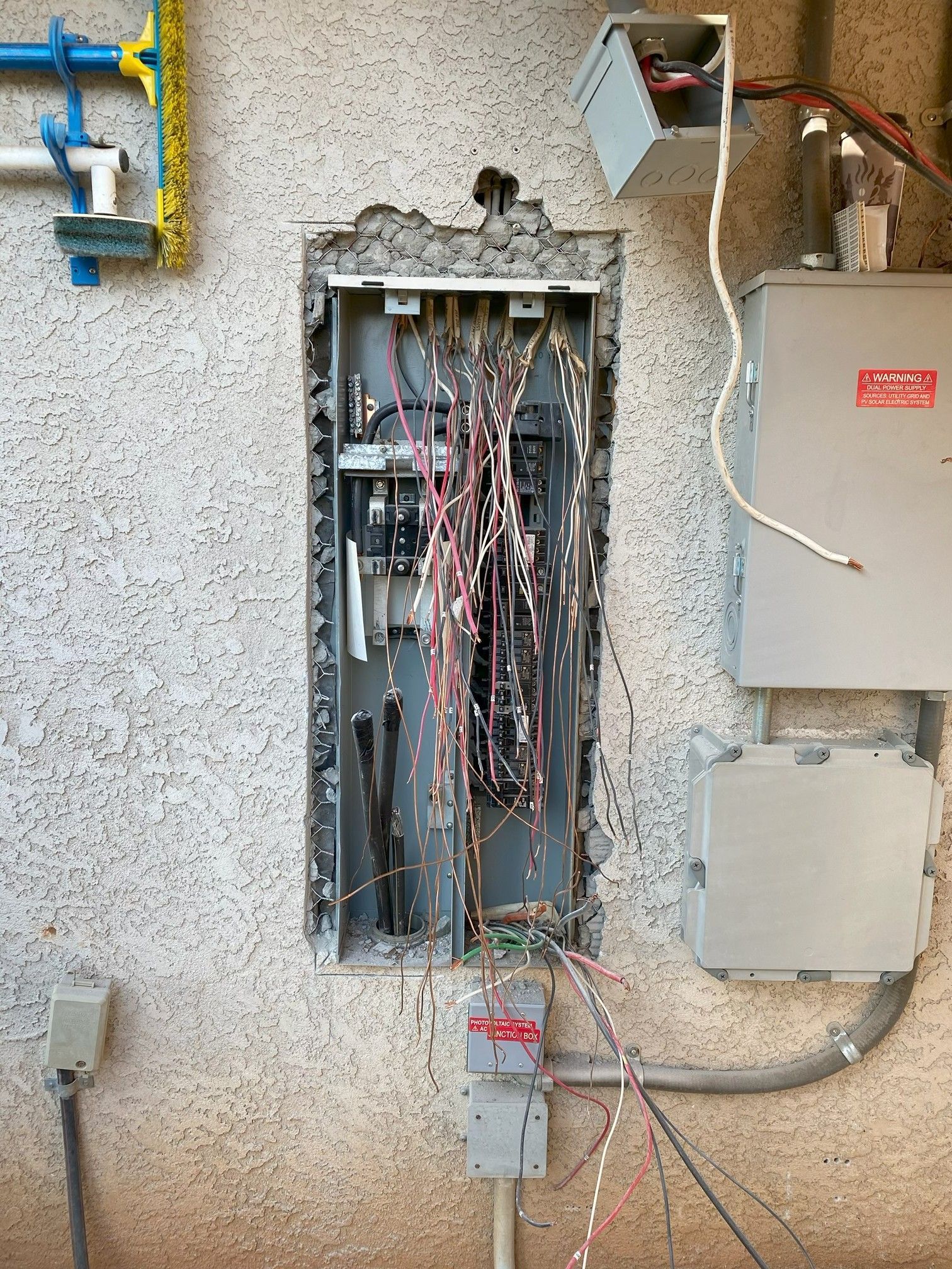 Open electrical panel with exposed wiring on a stucco wall and utility boxes nearby