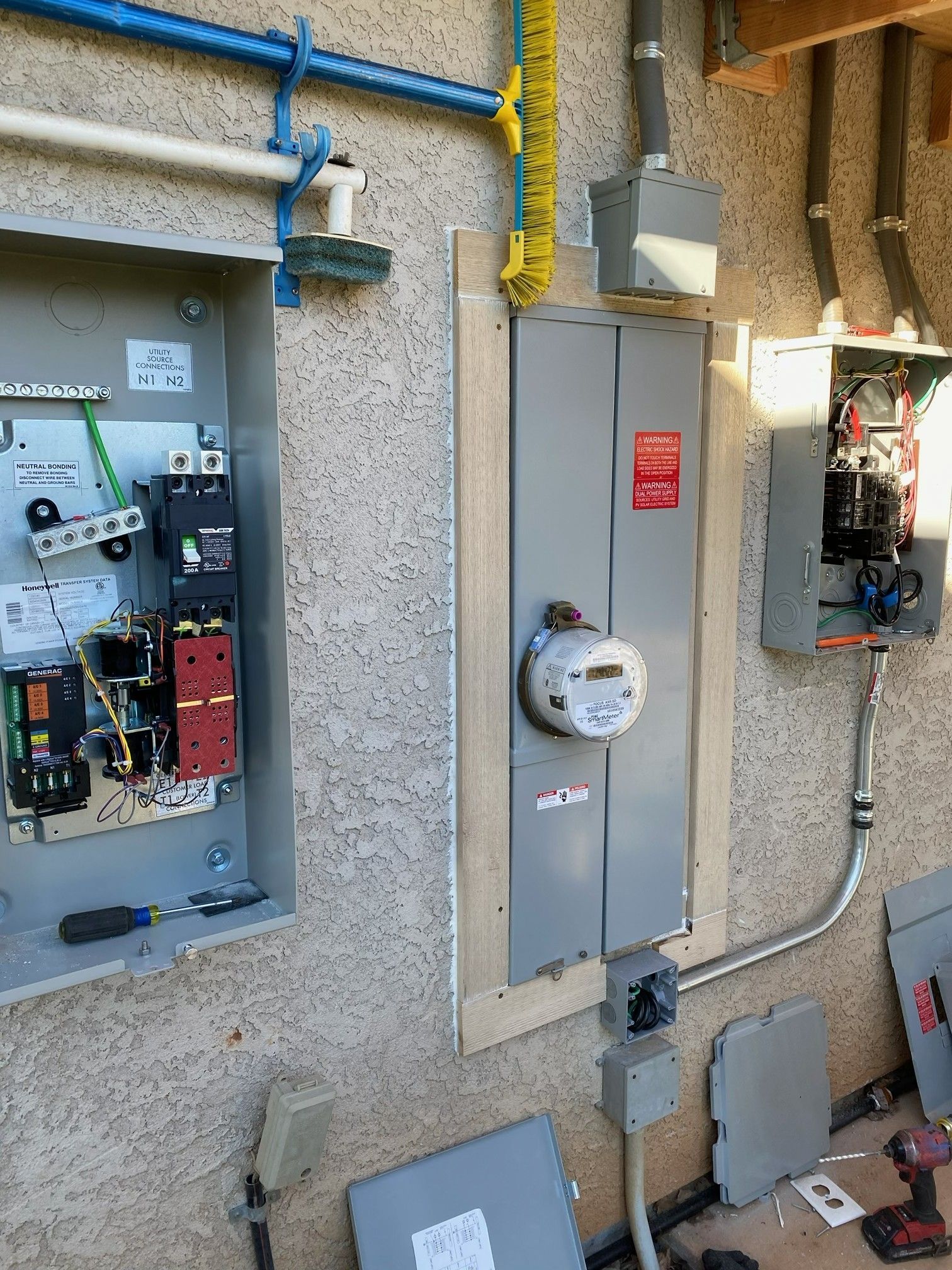 Electrical meter and breaker panels mounted on an exterior stucco wall with exposed conduit and pipes.