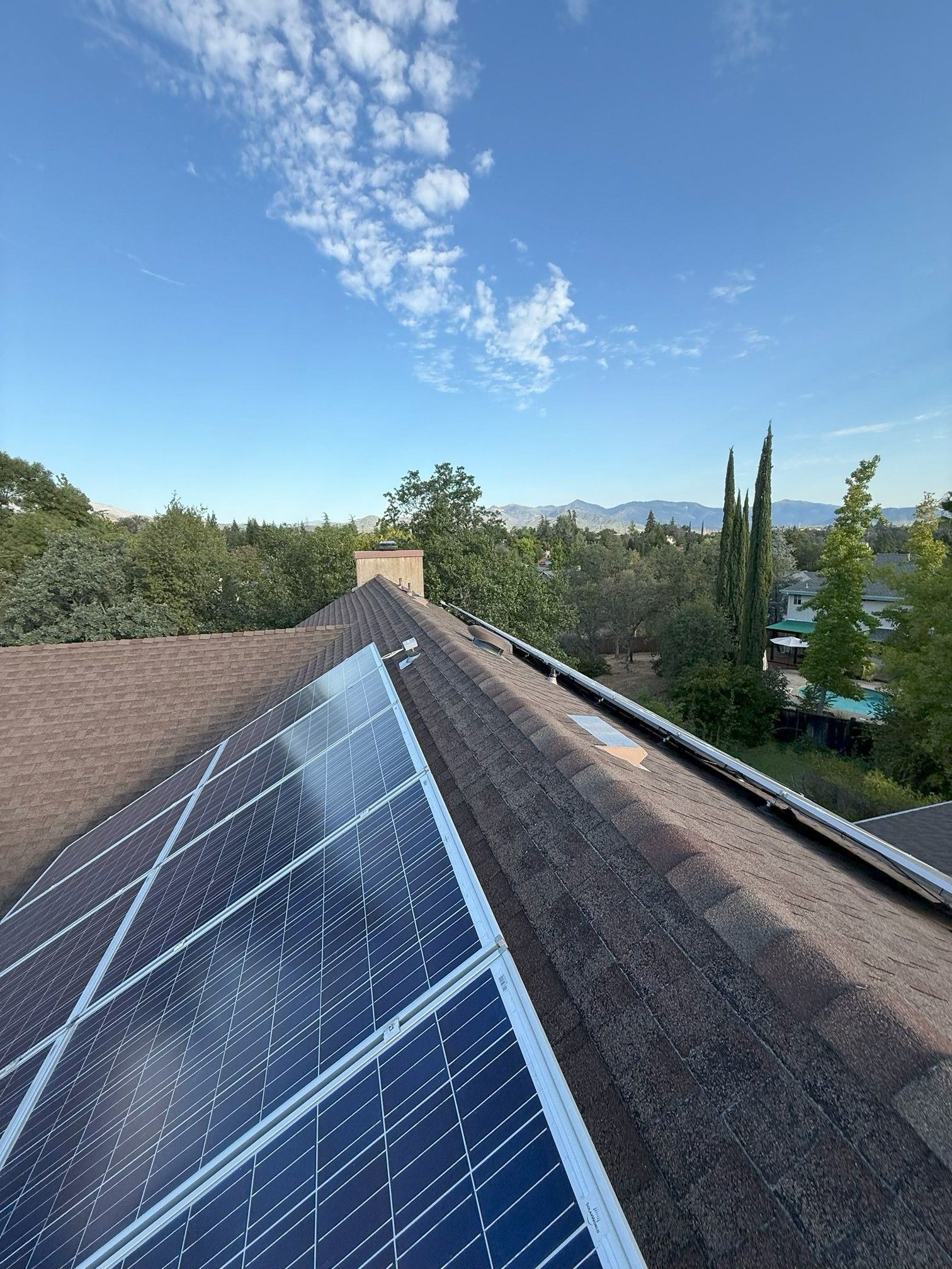Solar panels on a shingled rooftop under a clear blue sky, with trees and hills in the background.
