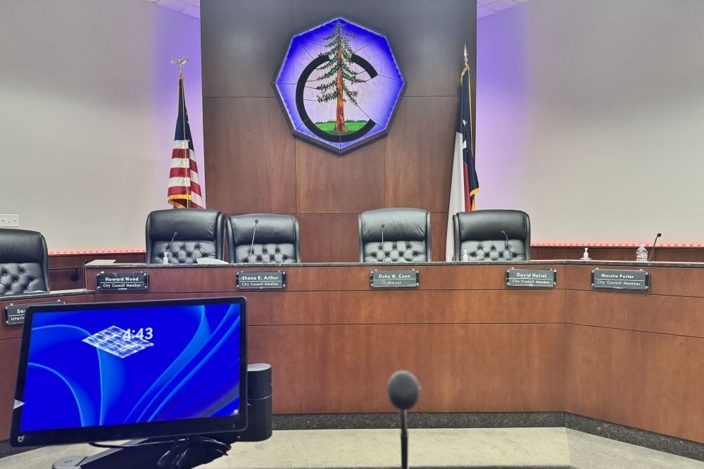 Empty council chamber with wood dais, five chairs, flags, and a wall seal behind it.