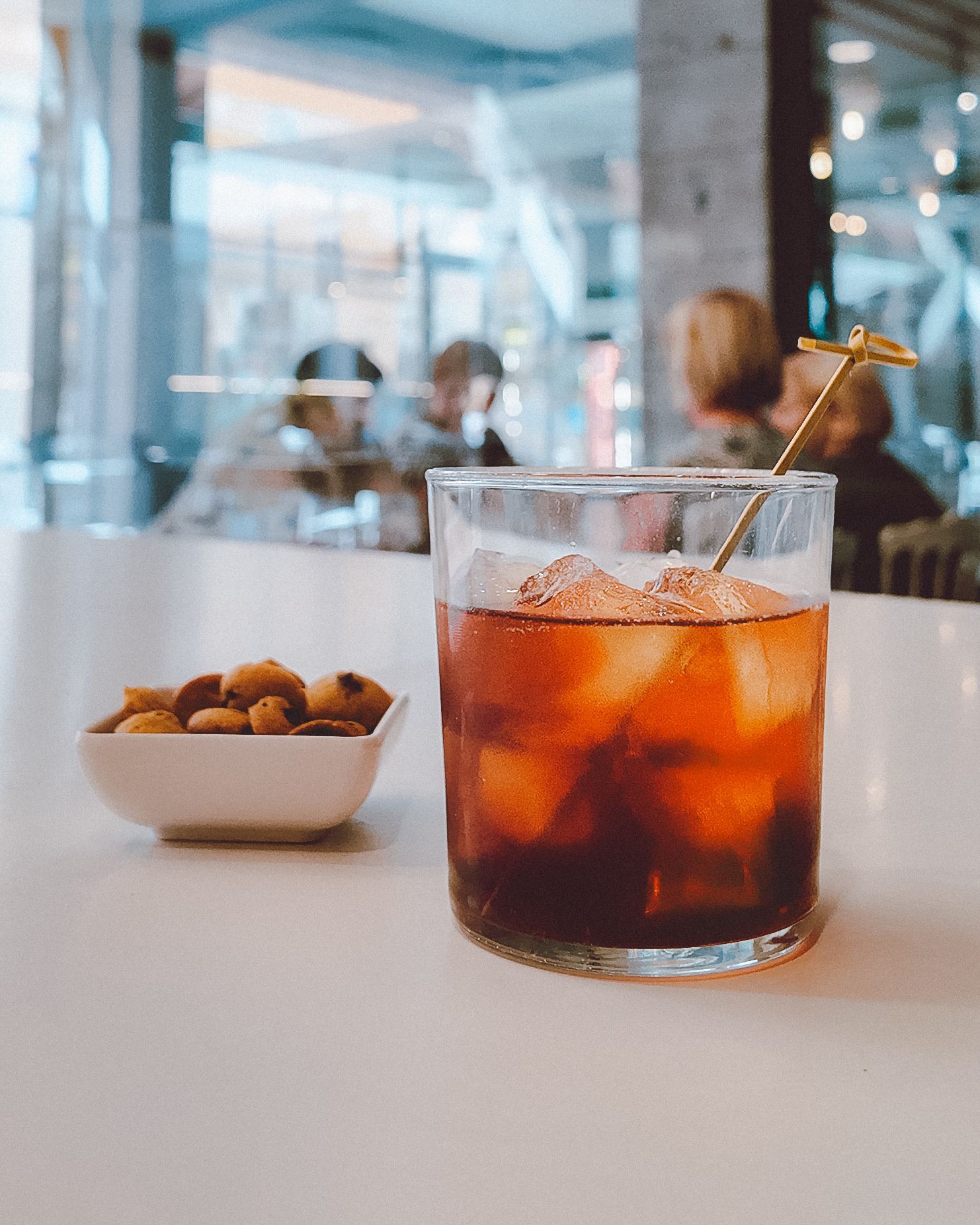 In un caffè, un cocktail ambrato ghiacciato con una guarnizione su un tavolo bianco accanto a una ciotola di noci.