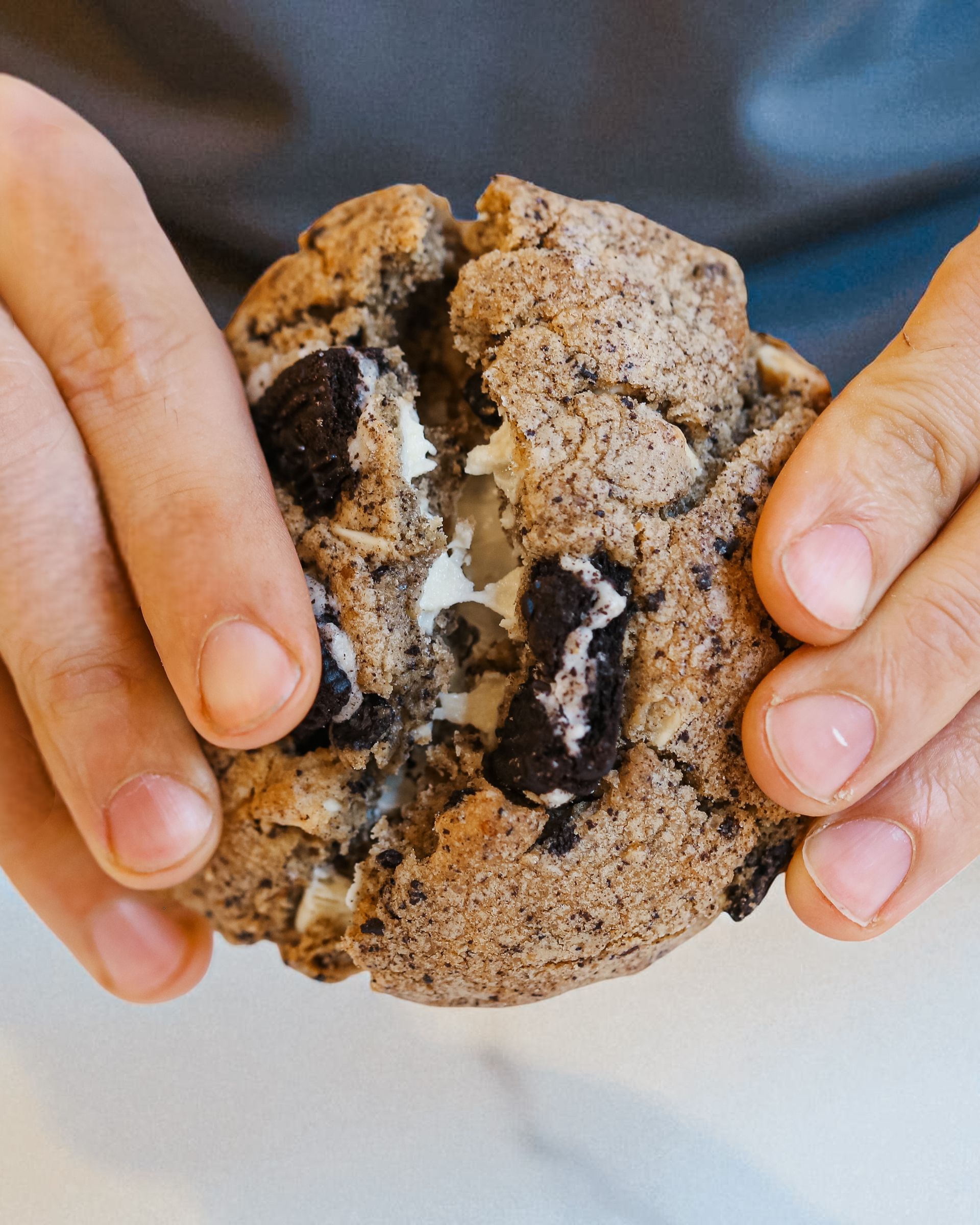 Mani che reggono un biscotto al cioccolato rotto con ripieno bianco su una superficie chiara
