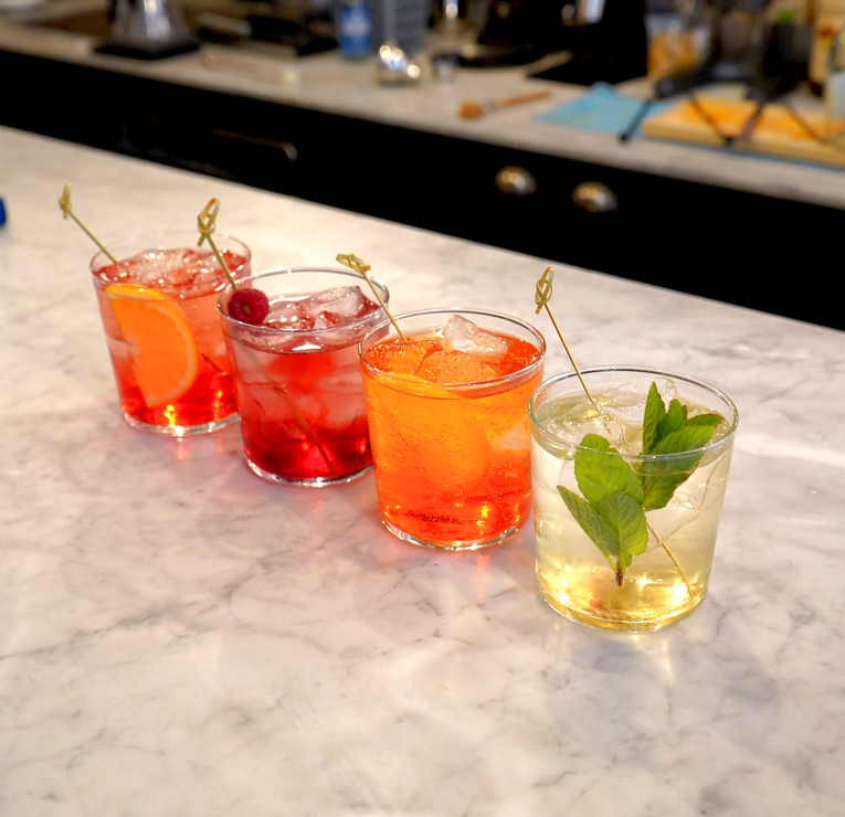 Quattro cocktail colorati in bicchieri bassi allineati su un bancone di marmo.
