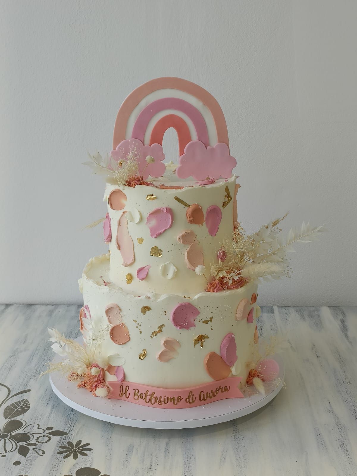 Torta a due piani rosa e crema con decorazione a forma di arcobaleno, fiori e la scritta