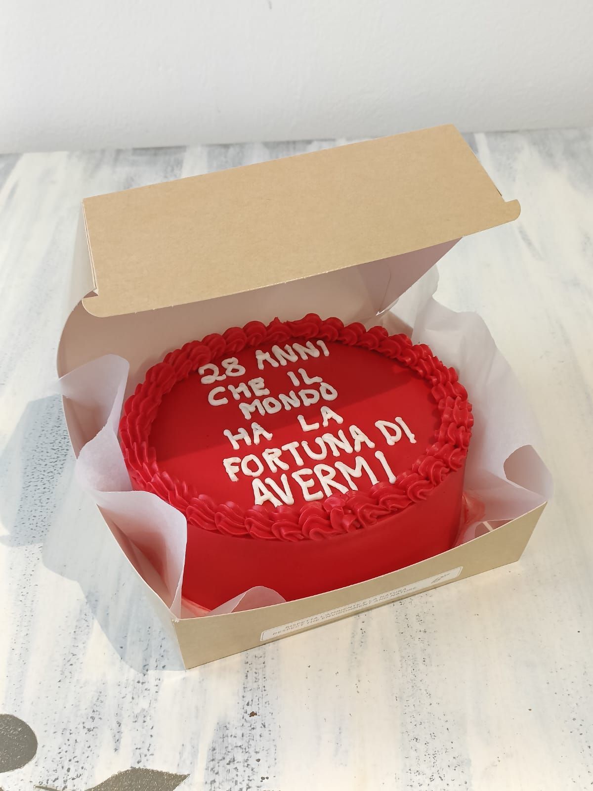 Torta rossa in una scatola aperta con scritta in glassa bianca, per celebrare un 28° anniversario in un'ambientazione floreale.