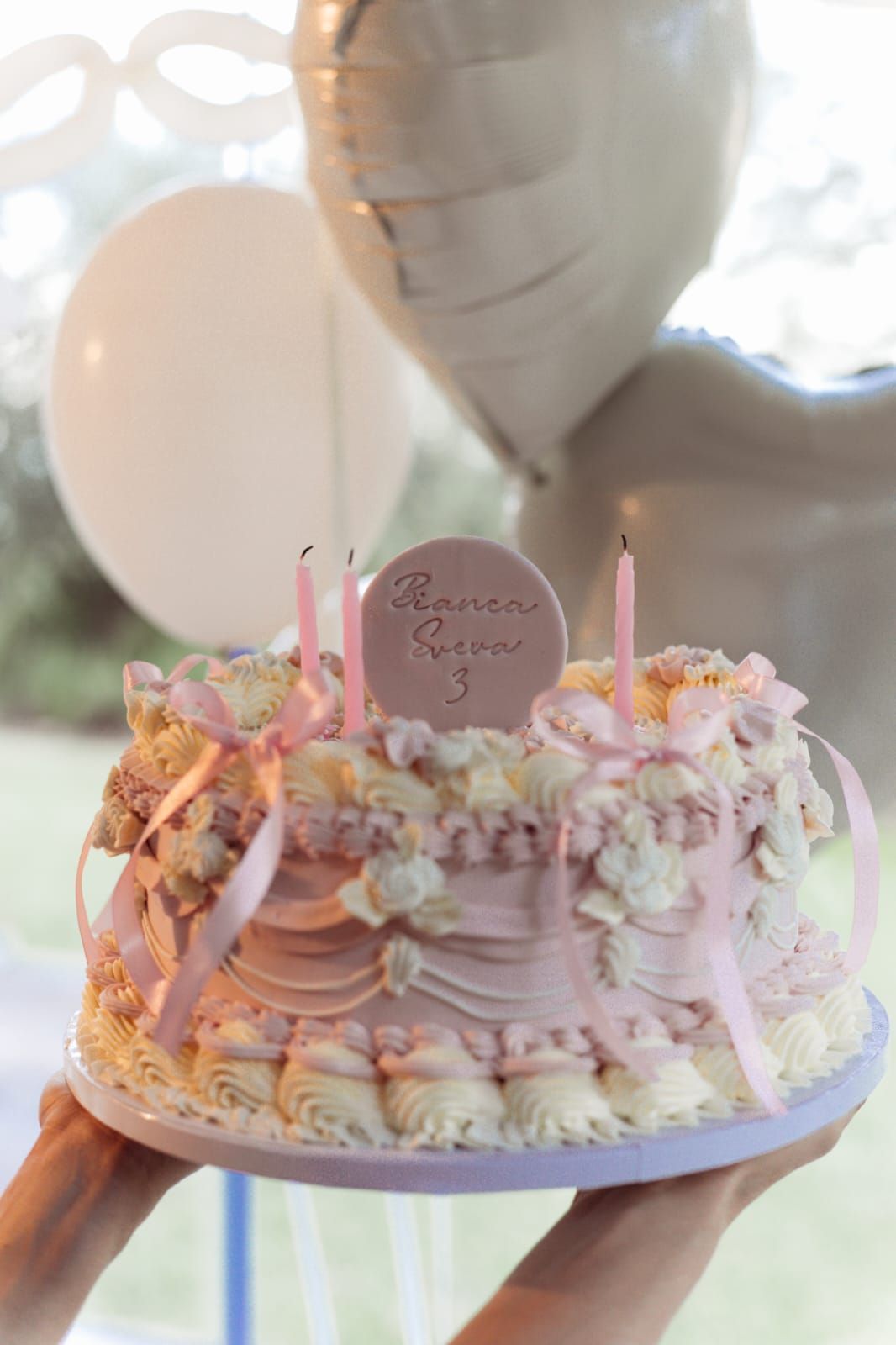 Torta di compleanno floreale rosa pastello con candeline e palloncini a forma di cuore sullo sfondo.