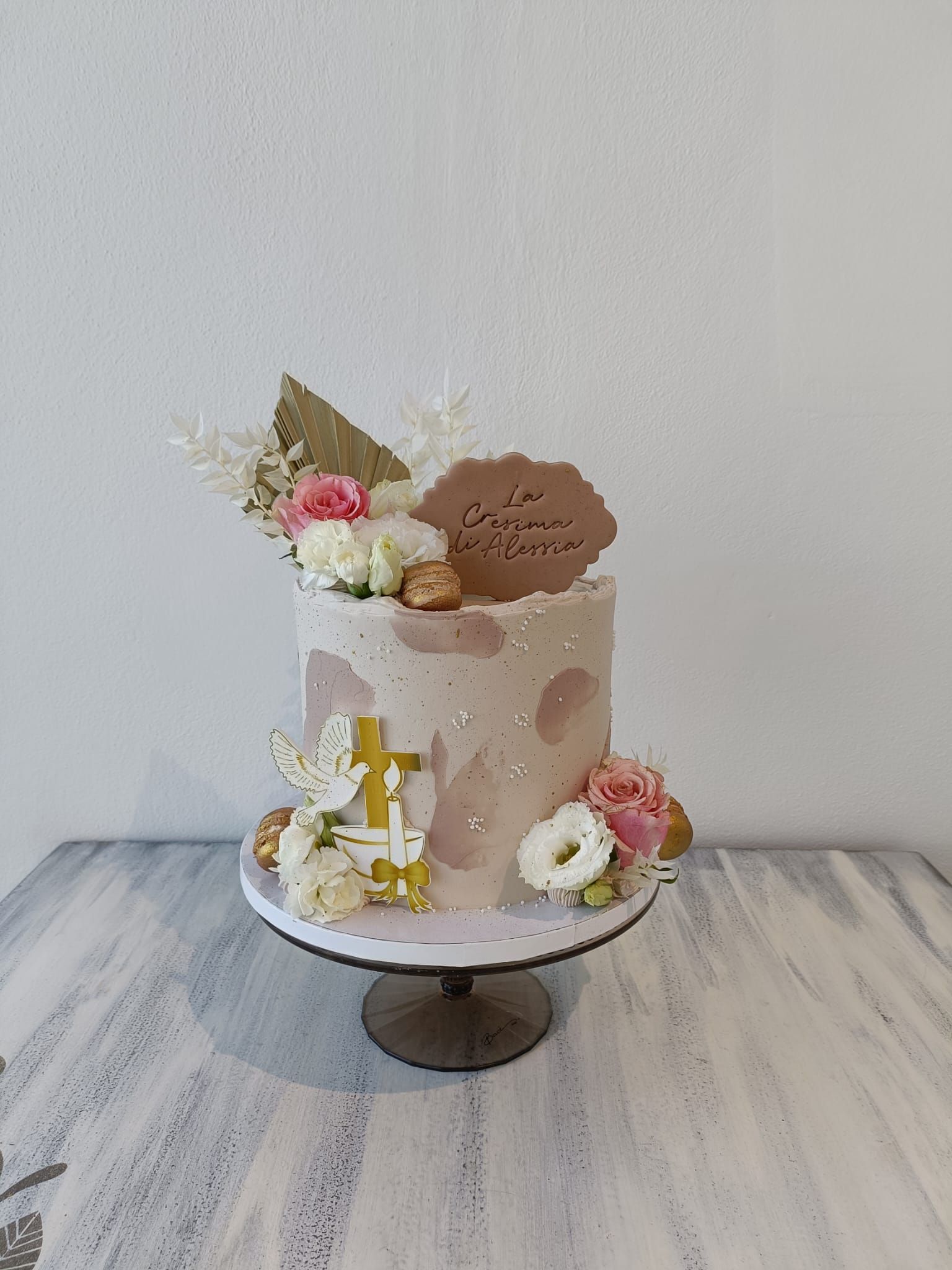 Torta decorata con colori pastello, su un'alzatina, guarnita con fiori e foglia d'oro, sullo sfondo di una parete chiara.