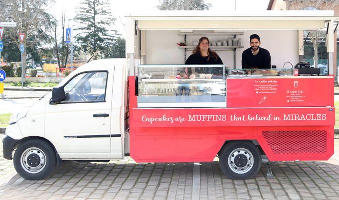 Un furgoncino rosso per la vendita di caffè, con due persone che servono i clienti dal finestrino in una strada cittadina.