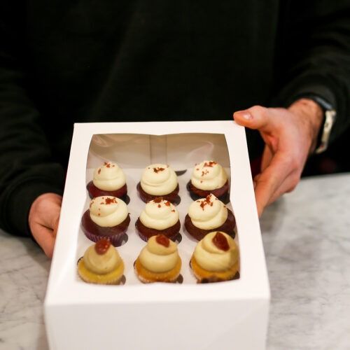 Mano che tiene una scatola bianca contenente nove mini cupcake con glassa bianca e decorazione rossa.