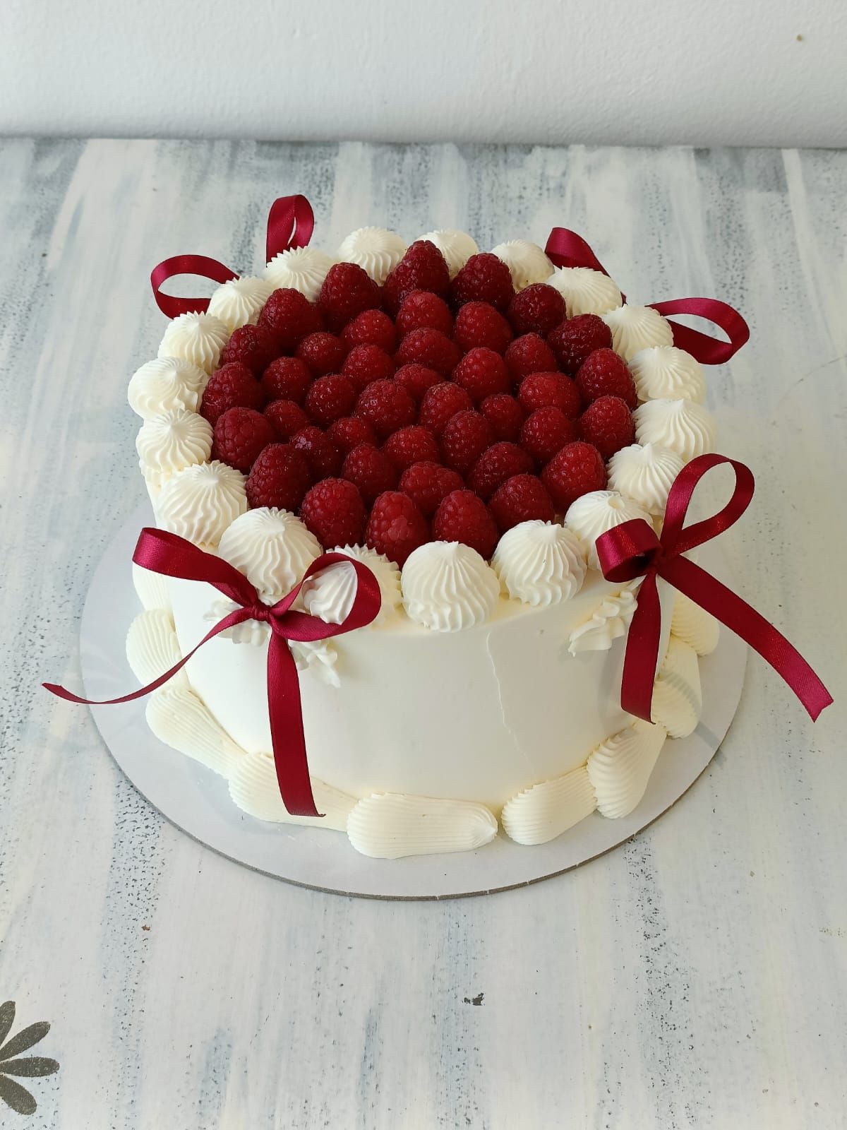 Torta bianca guarnita con lamponi rossi e decorata con fiocchi di nastro rosso su un tavolo bianco.