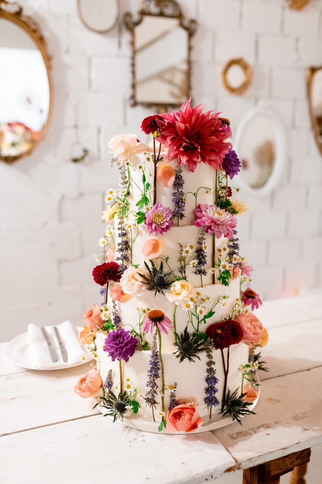 Torta floreale bianca a tre piani, decorata con fiori freschi colorati, su un tavolo rustico.