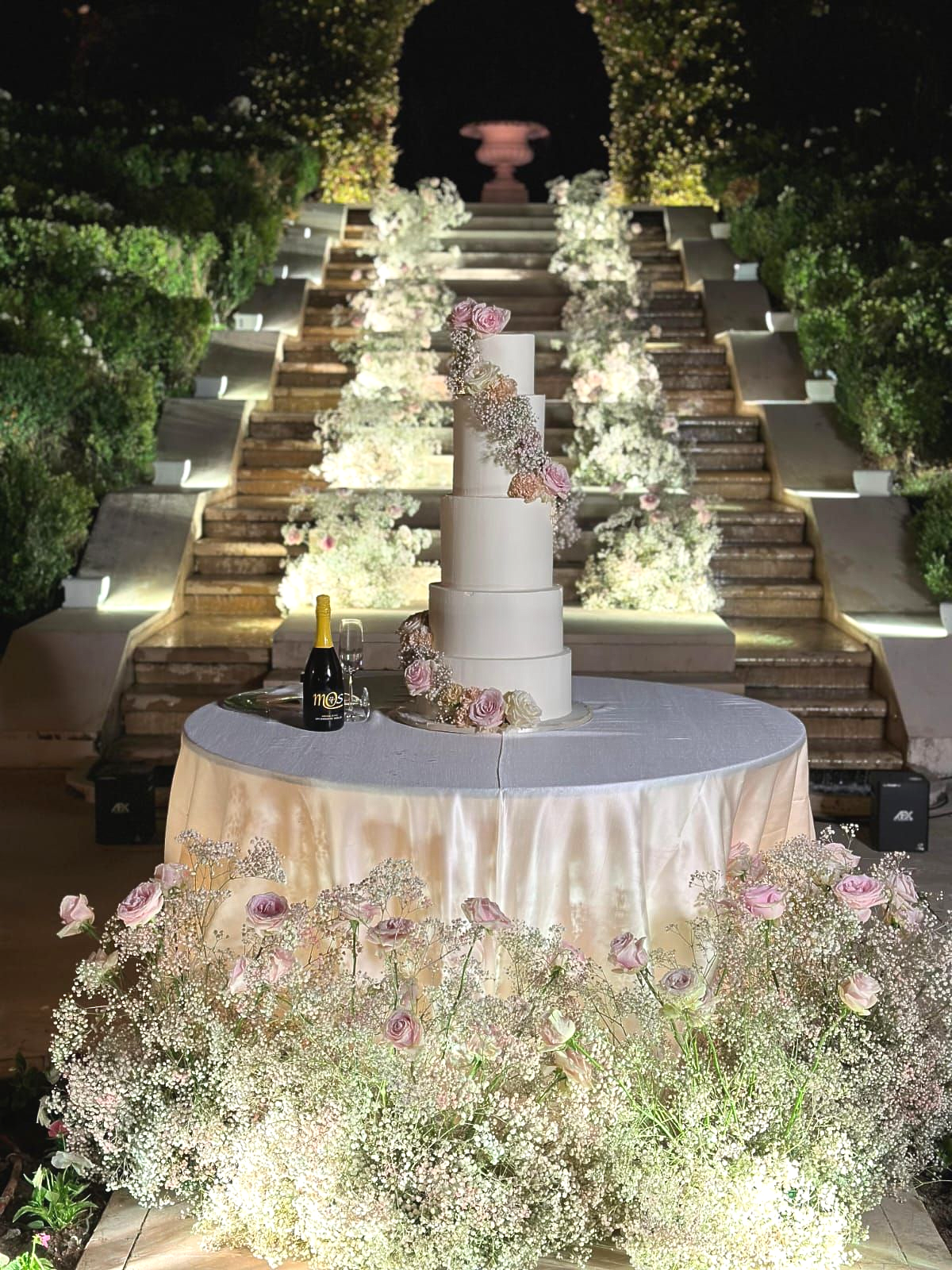 Una scalinata esterna fiancheggiata da fiori, illuminata da candele e con un tavolo rotondo come centrotavola di notte.