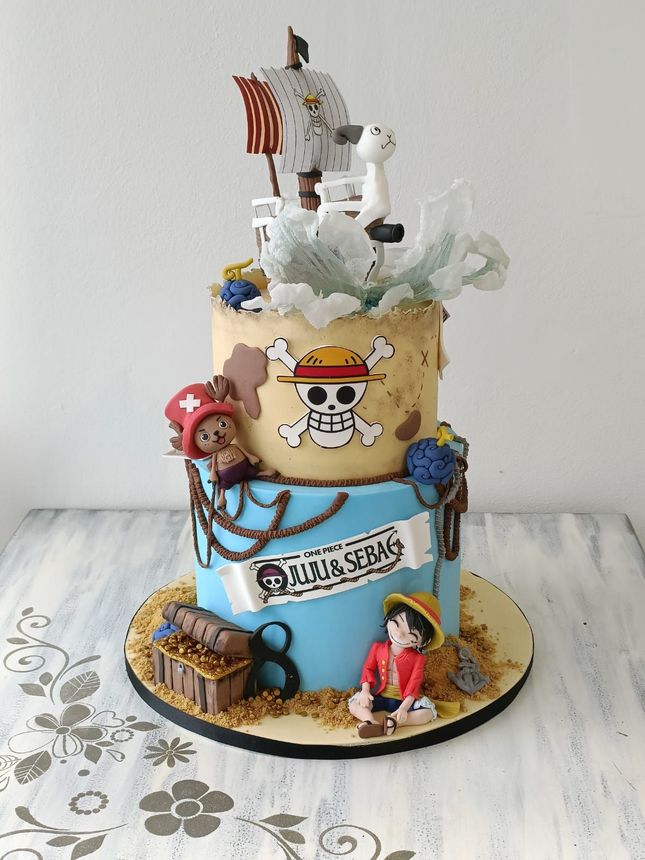 Torta a tre piani a tema One Piece con nave pirata, logo del teschio e piccole statuine dei personaggi sulla parte superiore e sulla base.