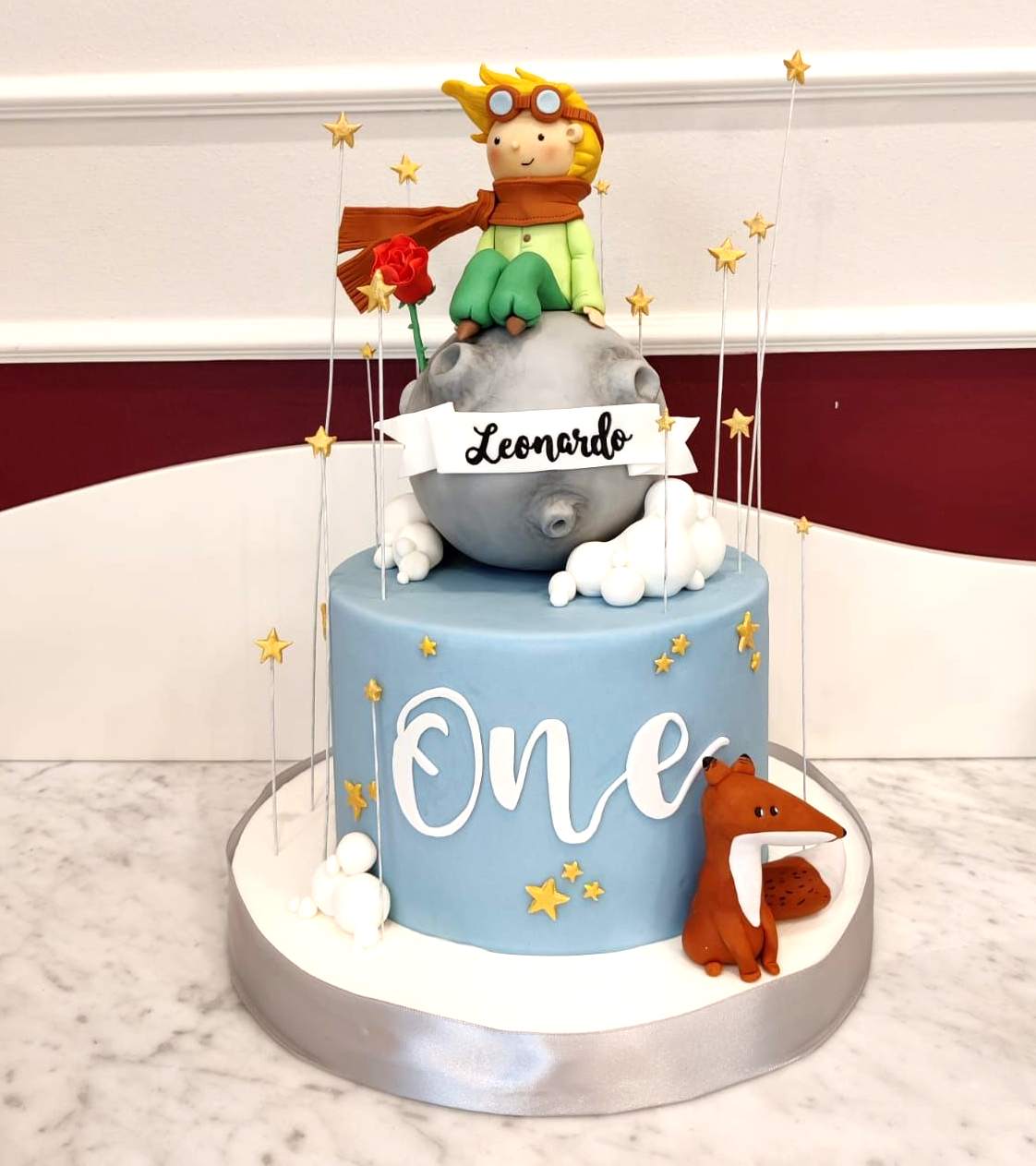 Torta di compleanno blu a un solo piano con una statuetta del Piccolo Principe, stelle, nuvole e una volpe su una base argentata.