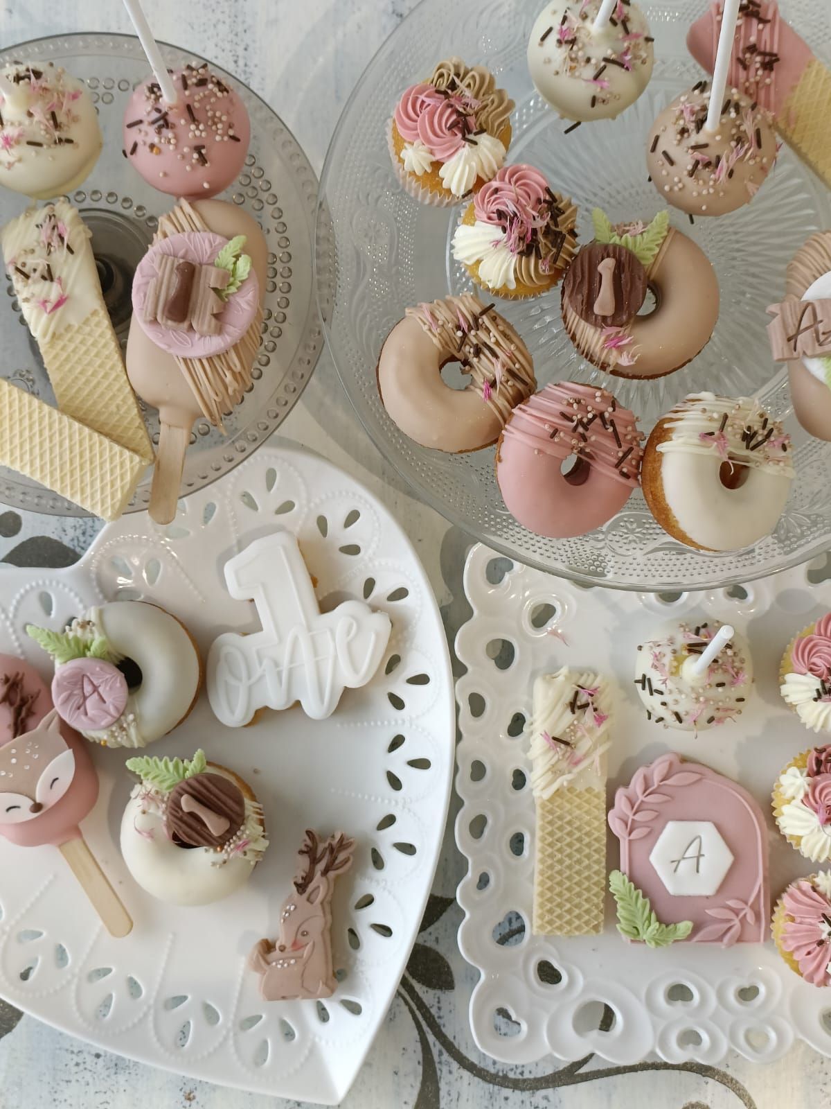 Assortimento di ciambelle e biscotti decorati con colori pastello su vassoi di pizzo, con caramelle e zuccherini.
