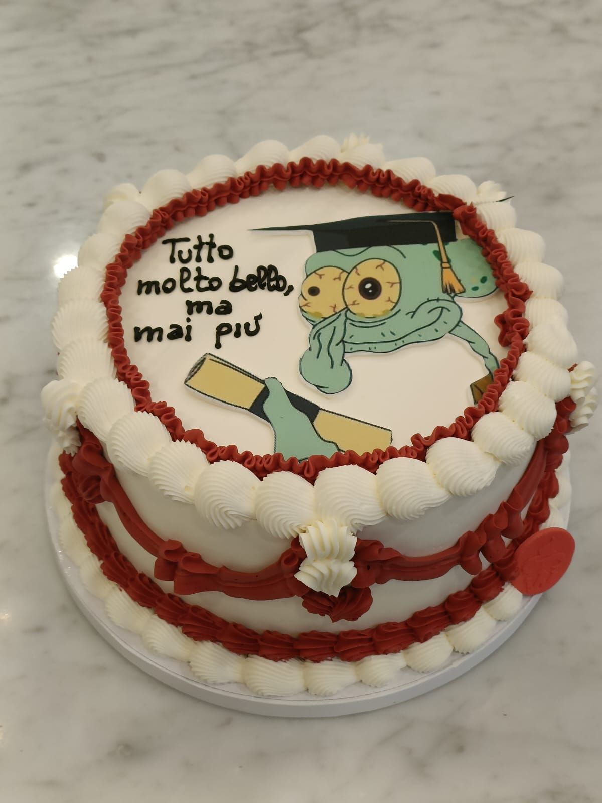 Torta rotonda bianca con glassa rossa, una statuina di laurea in stile cartone animato e la scritta in italiano