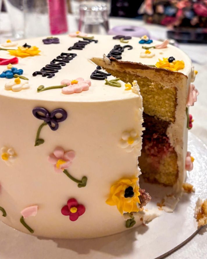 Torta di compleanno con glassa bianca, decorazioni floreali e una fetta mancante che lascia intravedere pan di Spagna giallo e ripieno di frutti di bosco.