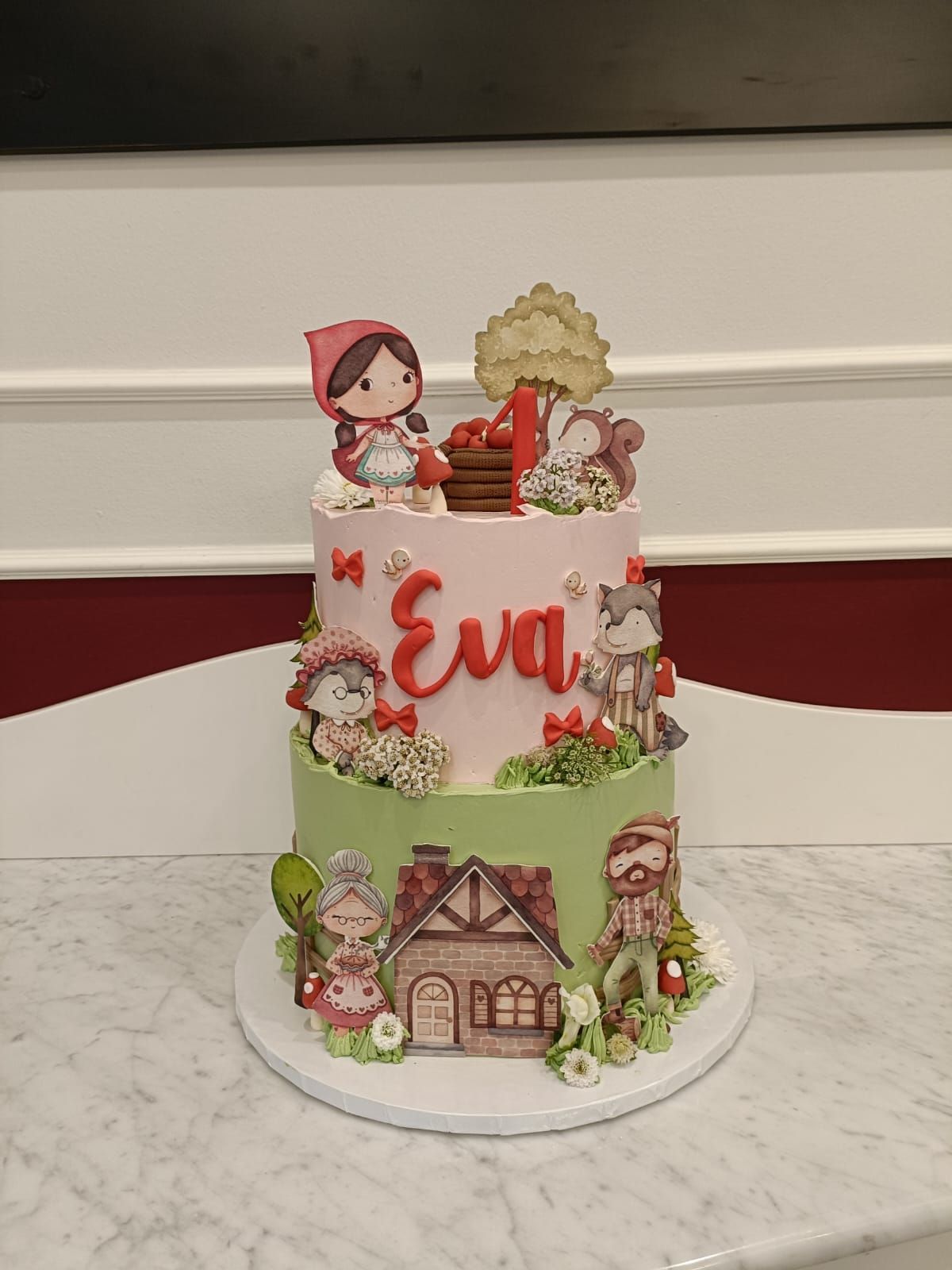 Torta di compleanno a due piani decorata con statuine di fatina e Cappuccetto Rosso, targhetta rossa con il nome e dettagli a forma di casa.