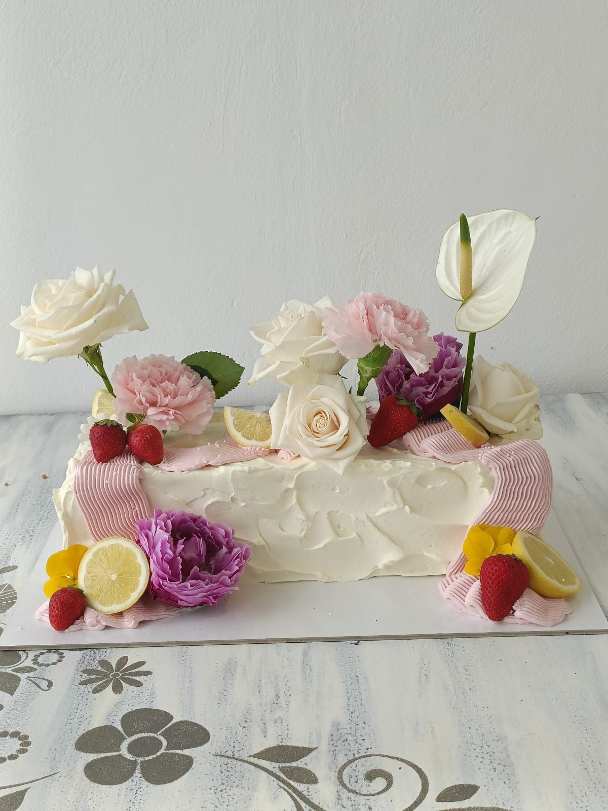 Torta bianca glassata, decorata con fiori, frutti di bosco, fette di limone e meringa, adagiata su una tovaglia.