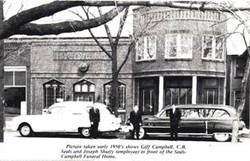 Historic black-and-white street scene with parked cars and a brick building under bare trees