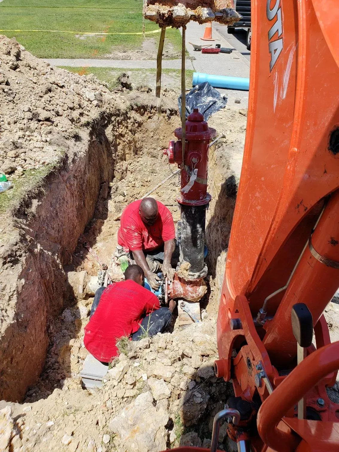 Two workers in red shirts repair a buried utility pipe in a dirt trench beside an excavator.