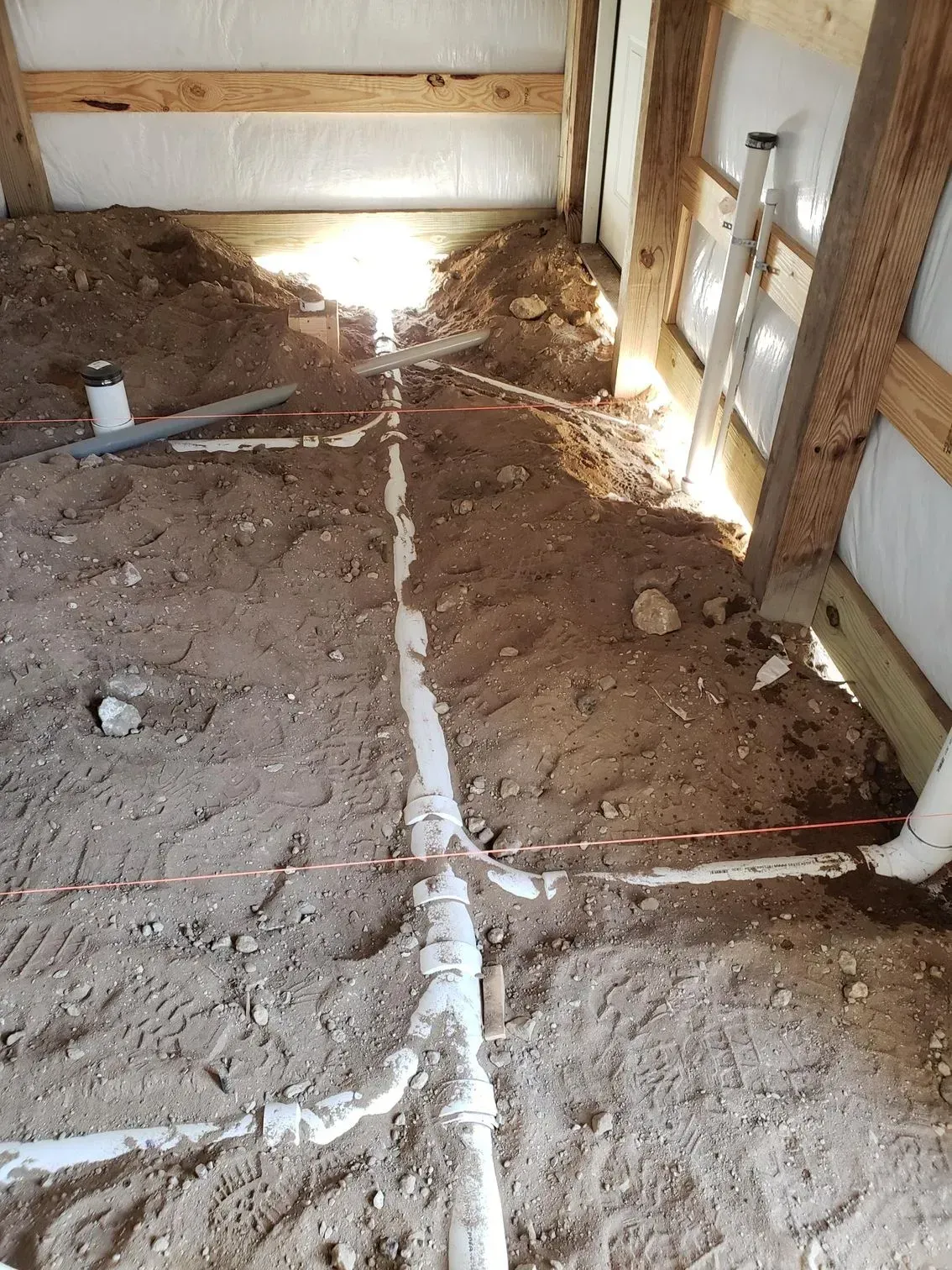 Dirt-floored unfinished room with white pipes and bright sunlight from an opening on the right.