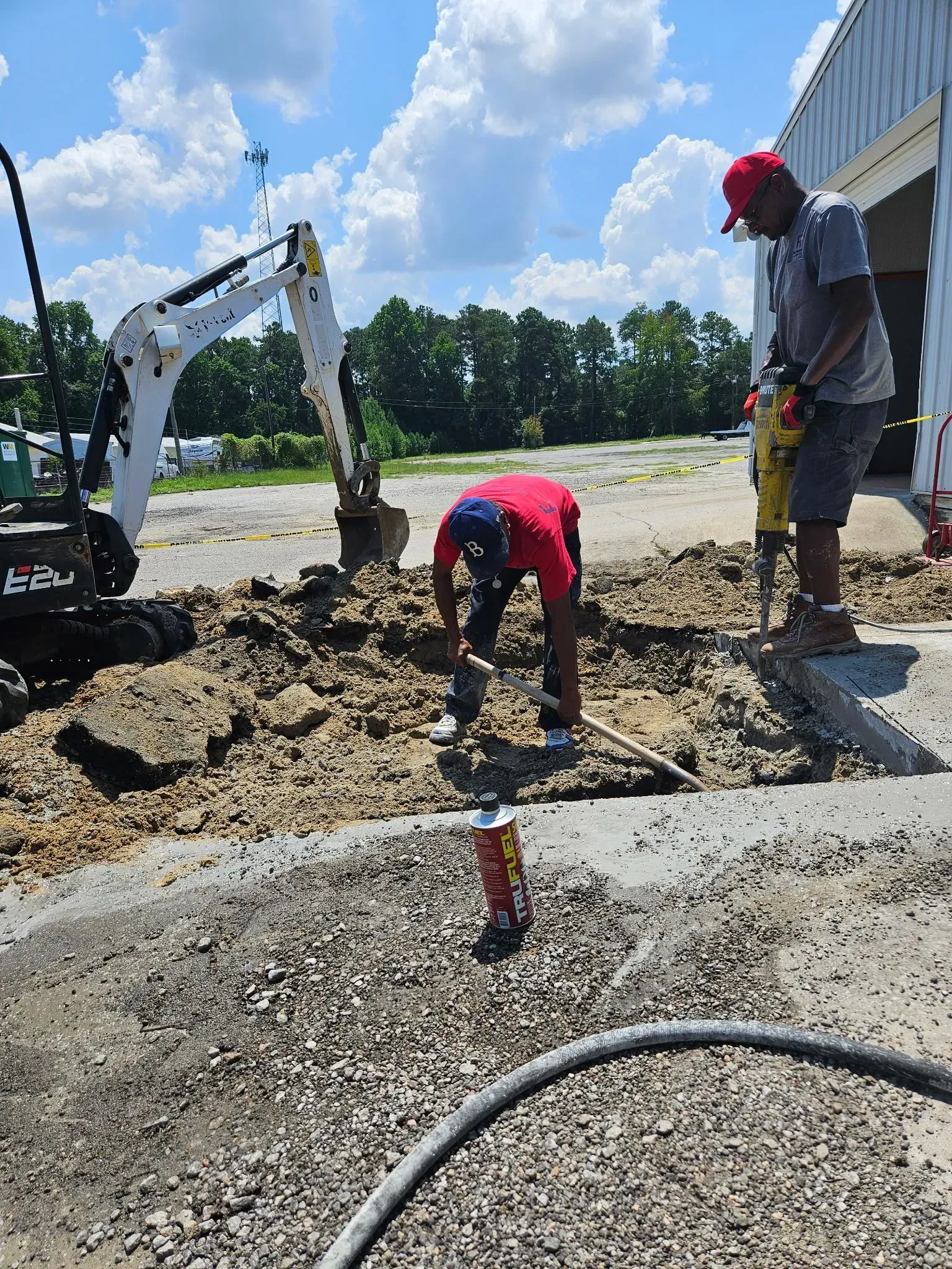Construction workers beside an excavator digging a trench on a gravel lot under a blue sky