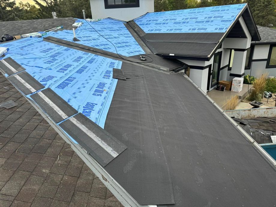 Roof under construction with blue underlayment and dark shingles on a sloped house roof