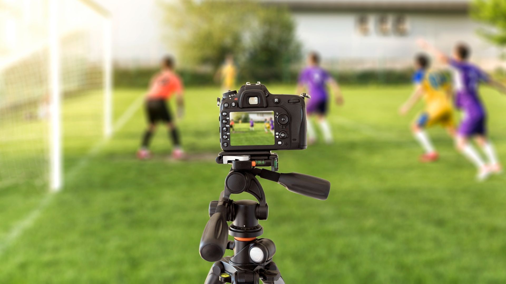 A camera on a tripod faces a soccer field, capturing a blurred view of players during a game near a goalpost.