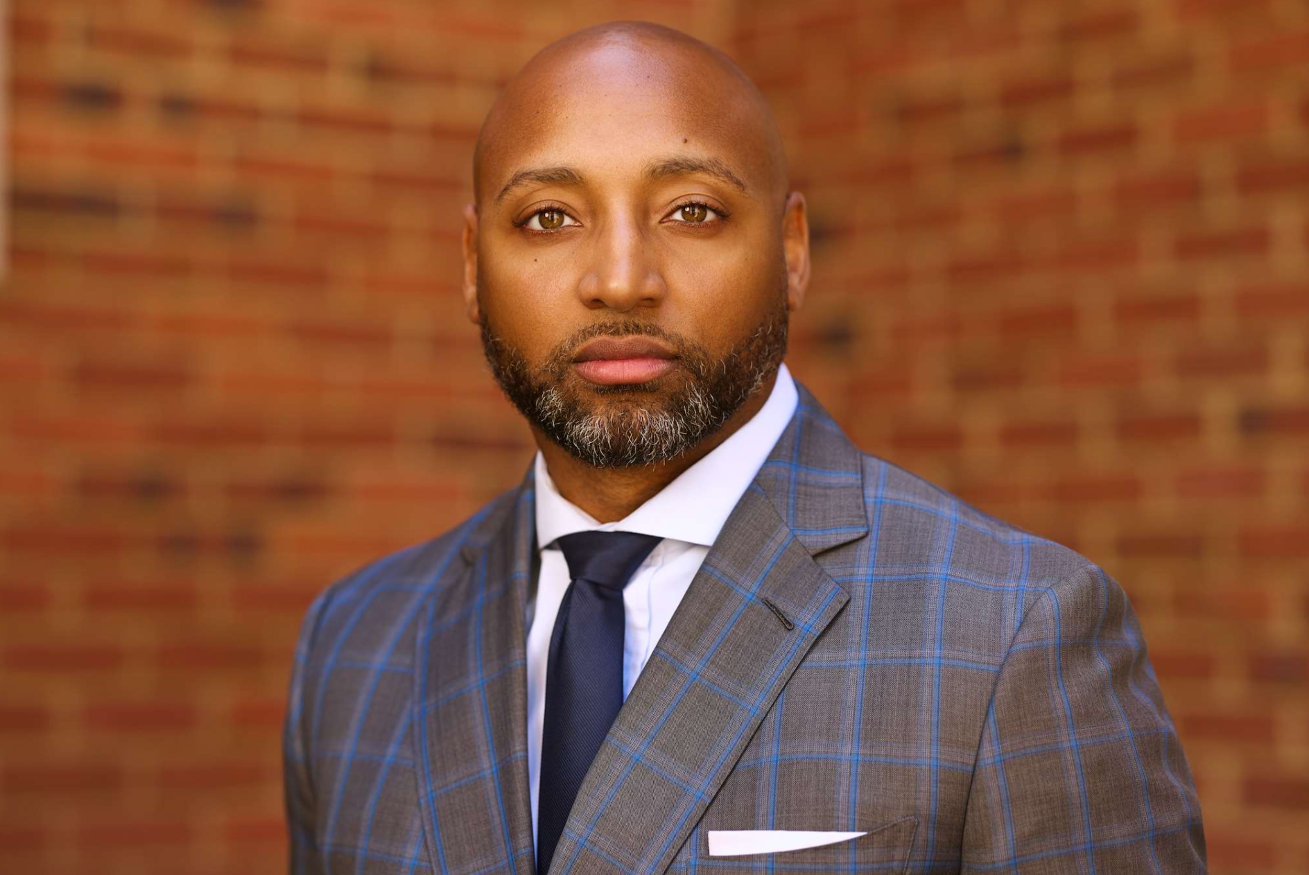 A person in a blue plaid double-breasted suit and light shirt, standing with hands clasped against a wood-slat background.