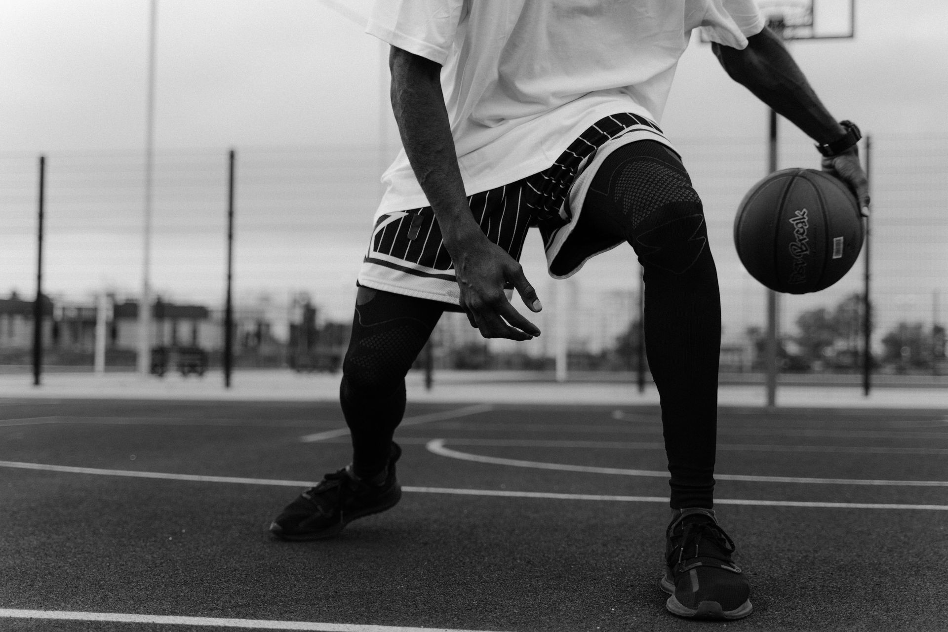A basketball player dribbling a ball on an outdoor court, wearing a t-shirt, athletic shorts, and compression leggings.