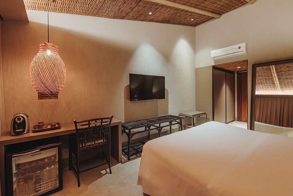 A modern hotel room with a desk, mini-fridge, black chair, mounted television, and a large bed under a thatched ceiling.