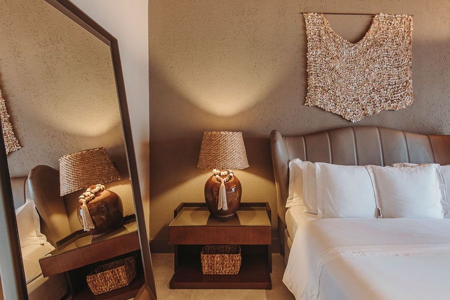 Um quarto com uma parede texturizada, uma mesa de cabeceira de madeira com um abajur, uma cama arrumada de branco e um espelho que reflete o ambiente.
