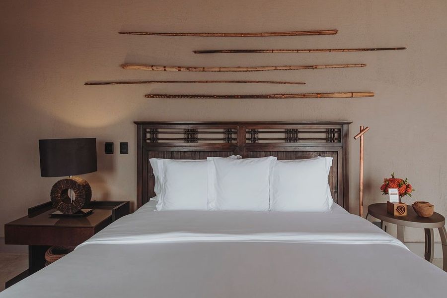 Um quarto com cabeceira de madeira escura, roupa de cama branca, duas mesas de cabeceira com abajures e obras de arte em madeira na parede.