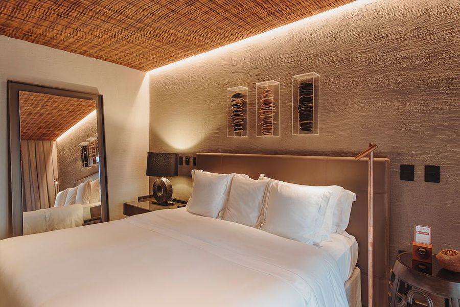 Um quarto moderno com teto texturizado em tecido trançado, um grande espelho de chão, uma cama em tons neutros e três obras de arte emolduradas na parede.