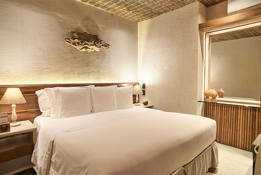 Um quarto de hotel com cama king-size, roupa de cama branca, cabeceira de madeira natural, iluminação ambiente e paredes texturizadas.