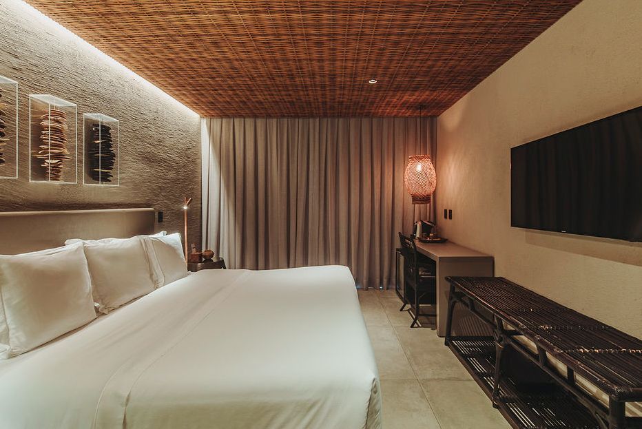 Modern hotel room with a large bed, textured ceiling, stone wall, desk, and flat-screen TV in warm, dim lighting.
