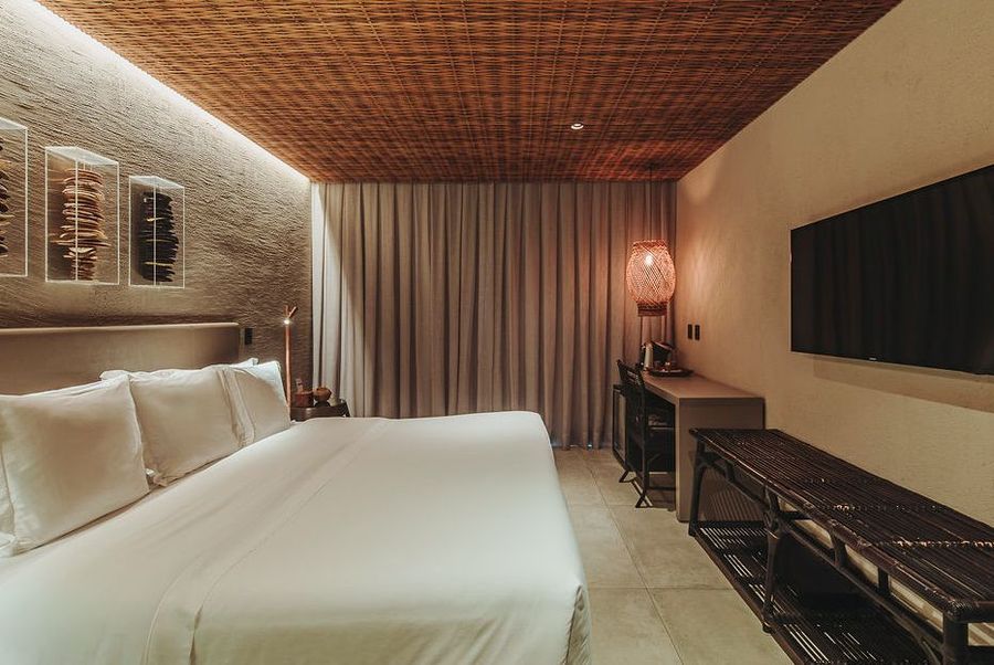 Um quarto de hotel moderno, em tons neutros, com cama branca, teto de madeira trançada, parede com detalhes em pedra e uma TV de tela plana fixada na parede.