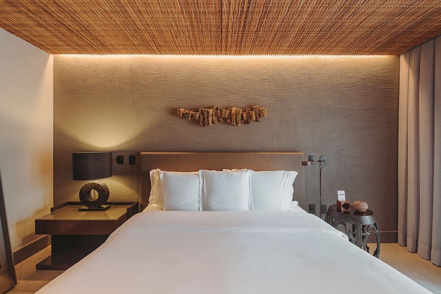 A bedroom featuring a queen bed with white linens, textured brown walls, a woven wood ceiling, and warm ambient lighting.